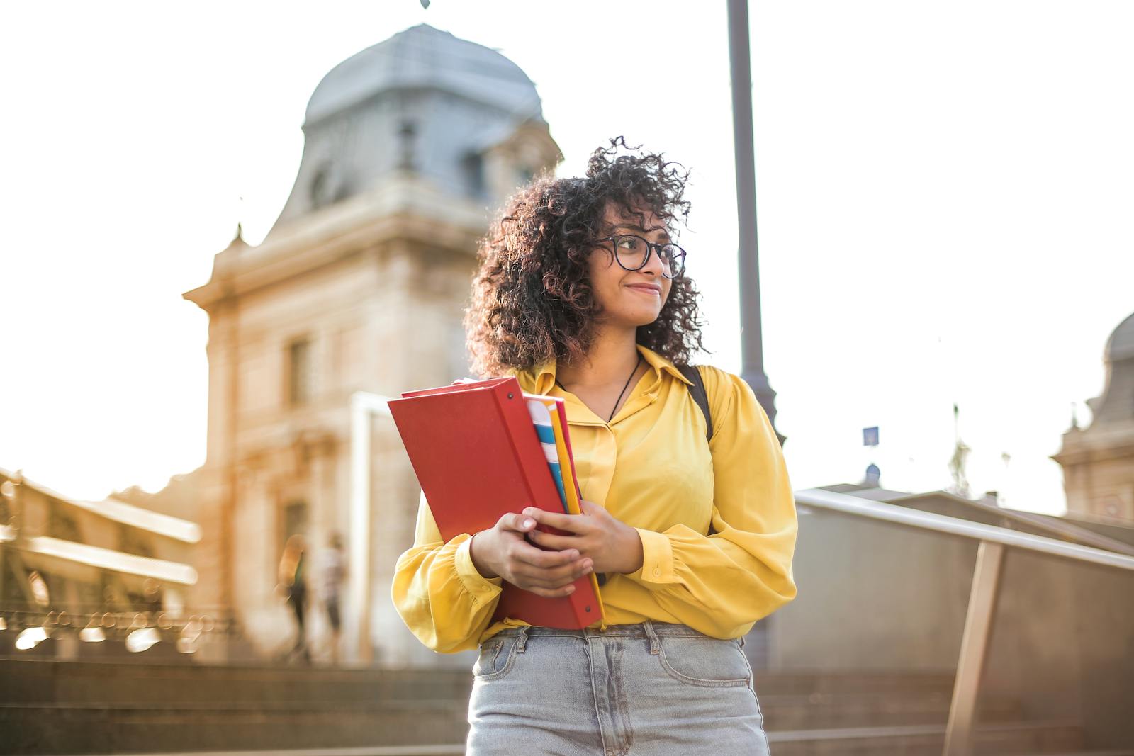 Photo by Andrea Piacquadio Smiling female student with curly hair and glasses holding books on campus steps.